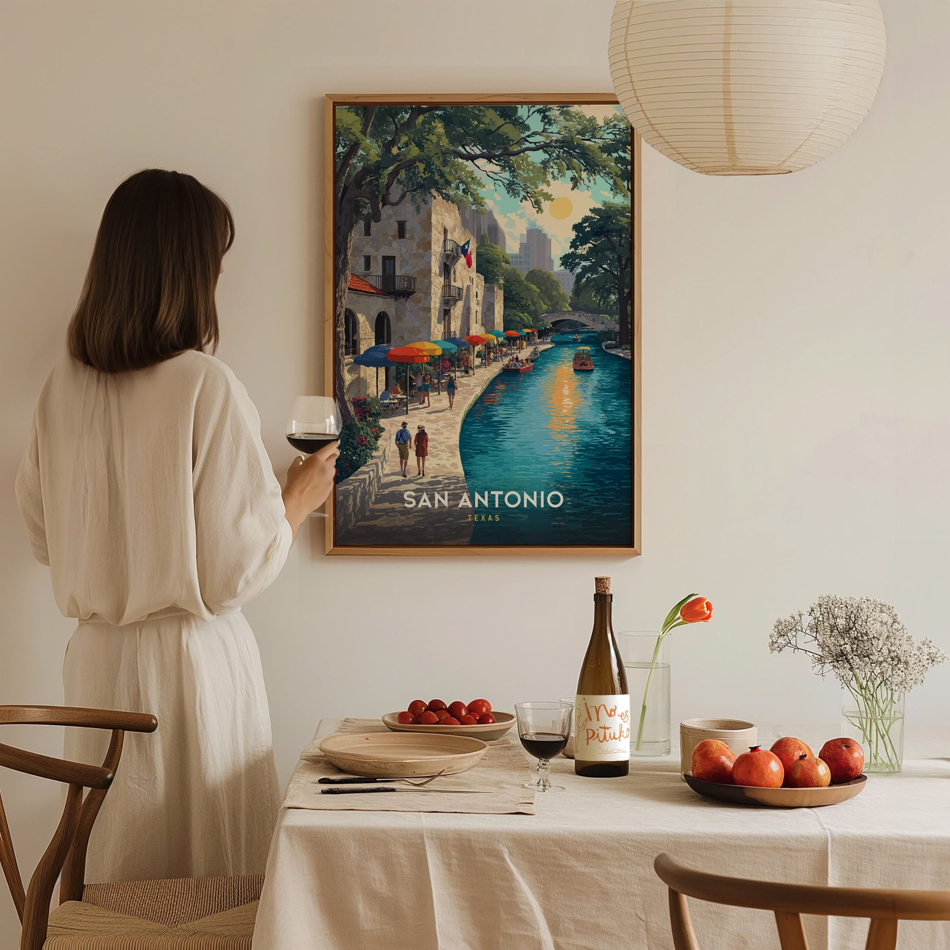 A woman in a white dress holds a glass of wine, admiring a colorful San Antonio Riverwalk poster on a wall above a dining table with fruit and wine.