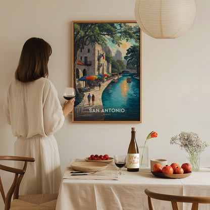 A woman in a white dress holds a glass of wine, admiring a colorful San Antonio Riverwalk poster on a wall above a dining table with fruit and wine.