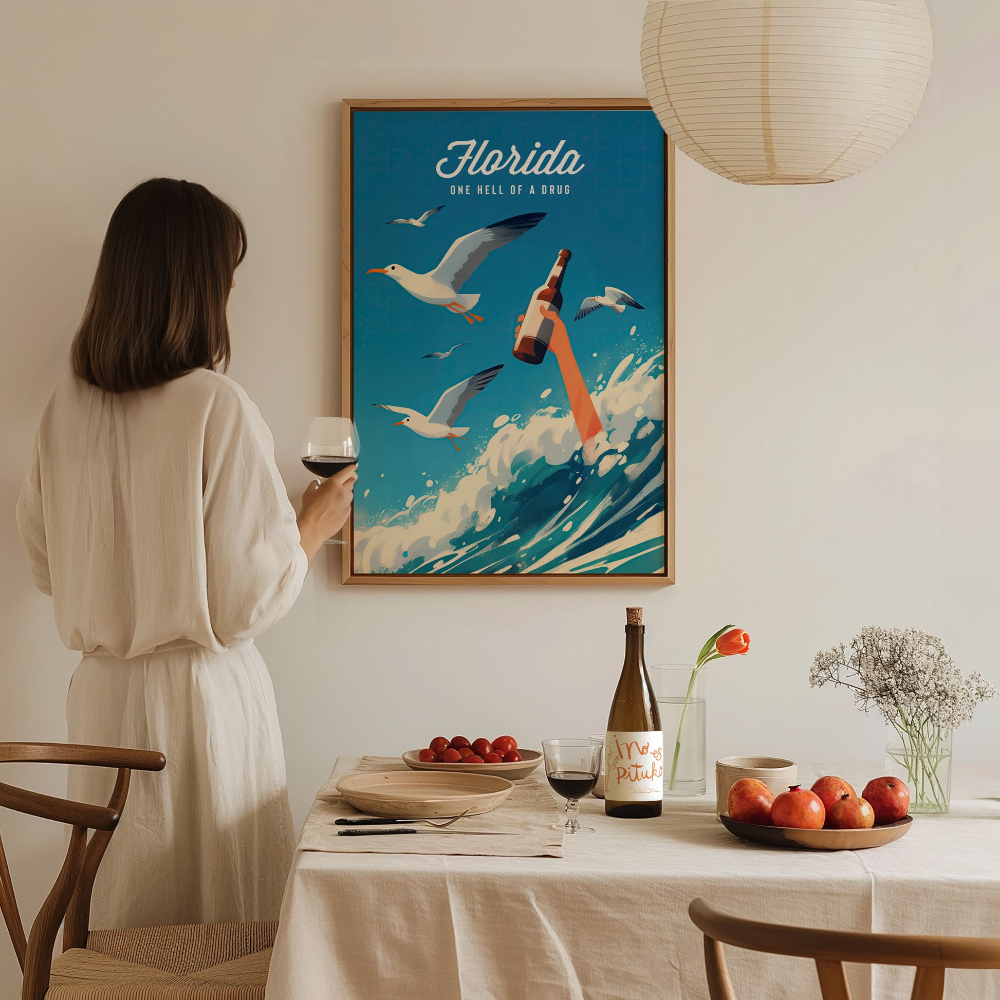 A woman in a white dress holds a wine glass, admiring a colorful poster of seagulls and a bottle among ocean waves.