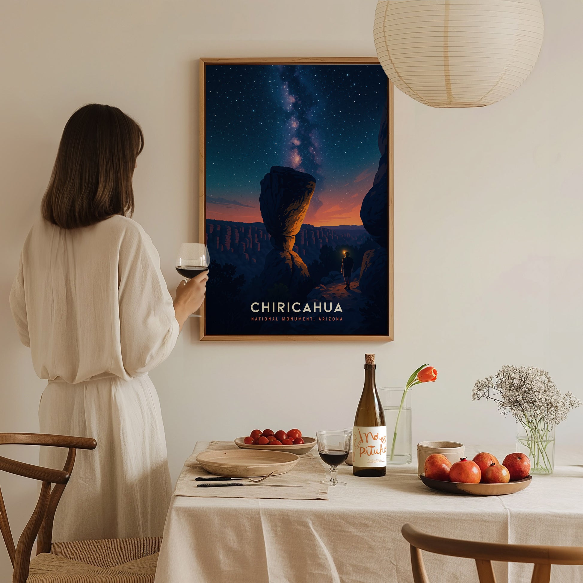 A woman in a white outfit stands holding a glass of wine, gazing at a framed poster of Chiricahua National Monument, featuring a starry sky above dramatic rock formations.