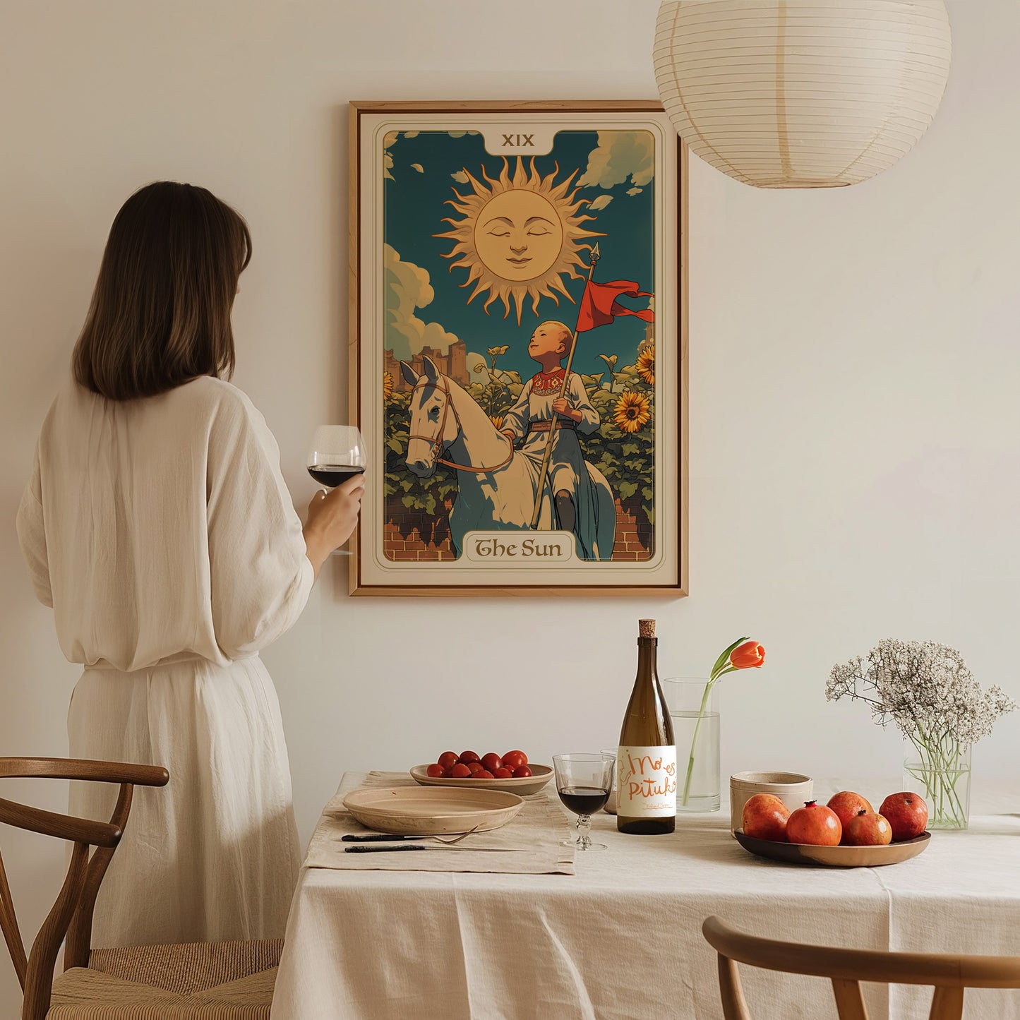 A woman in a white dress holds a glass of wine, admiring a framed Sun tarot card poster above a table with fruit, wine, and flowers.