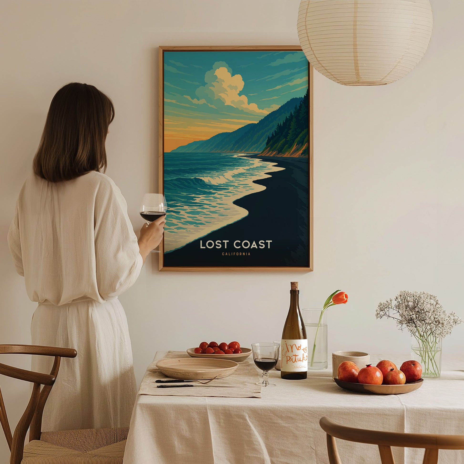 A woman in a light-colored outfit stands holding a glass of wine, admiring a colorful framed poster titled Lost Coast featuring a serene beach scene with waves and mountains.