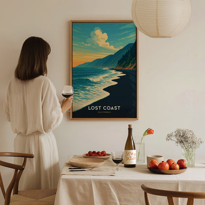 A woman in a light-colored outfit stands holding a glass of wine, admiring a colorful framed poster titled Lost Coast featuring a serene beach scene with waves and mountains.