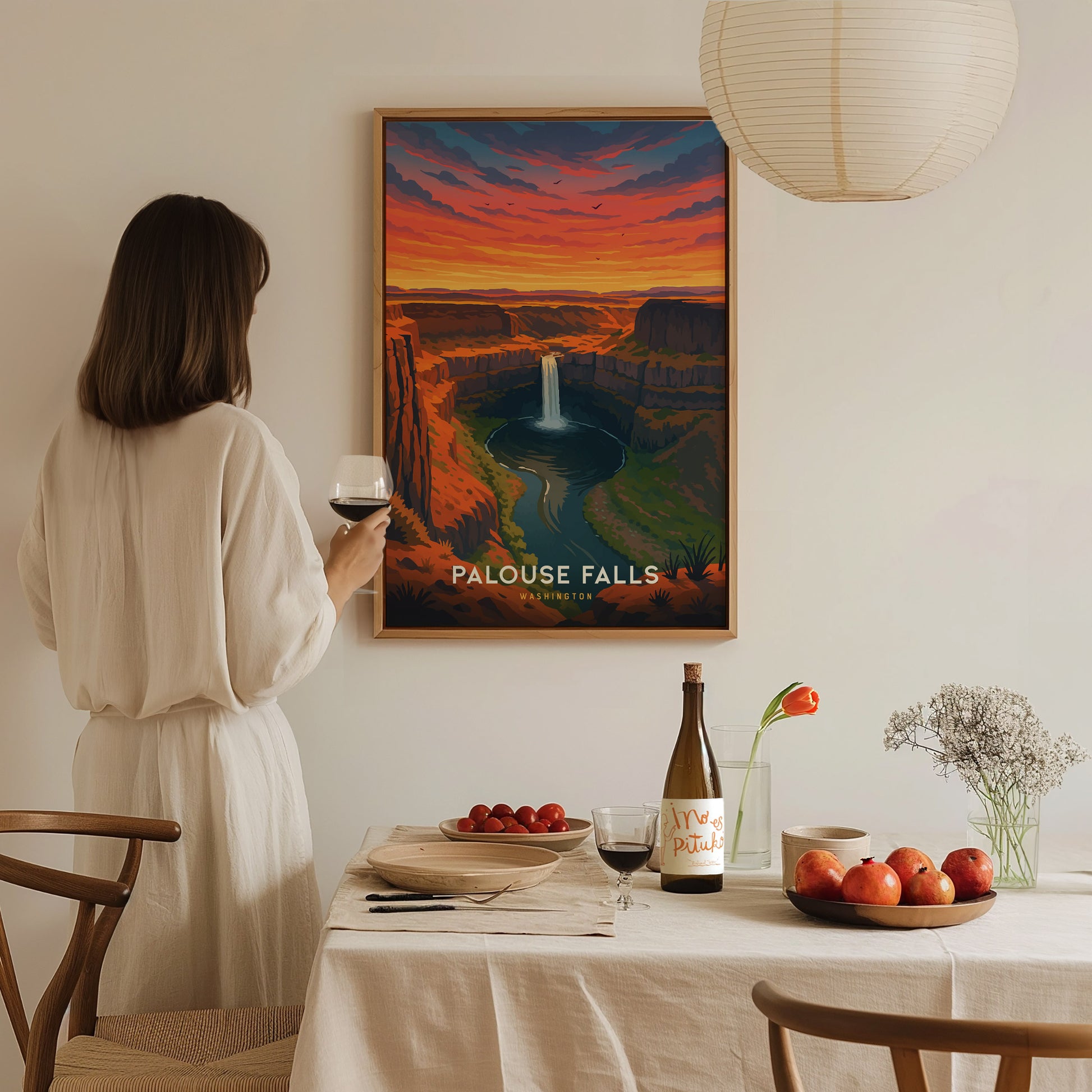 A person in a light-colored outfit stands in front of a vintage-style poster of Palouse Falls, showcasing a vibrant sunset over a canyon with a waterfall. The scene is set in a cozy dining area with a table adorned with fruit and a bottle.