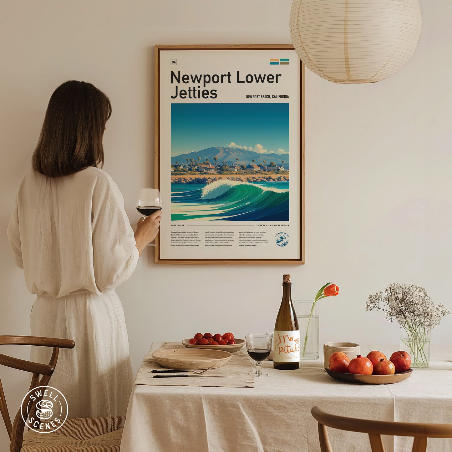 A woman in a white dress holds a glass of wine, admiring a framed Newport Lower Jetties poster on the wall.
