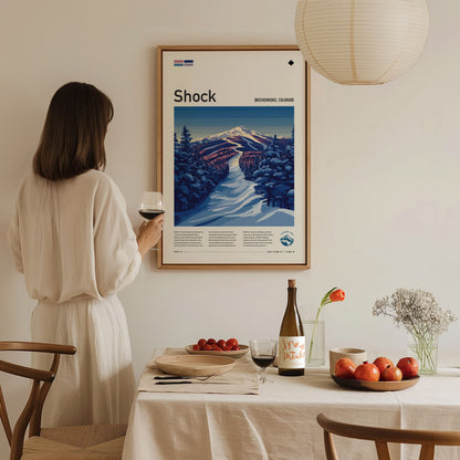 A woman in a white dress holds a glass of wine while admiring a framed ski run poster on the wall. A dining table with fruit and wine is in.