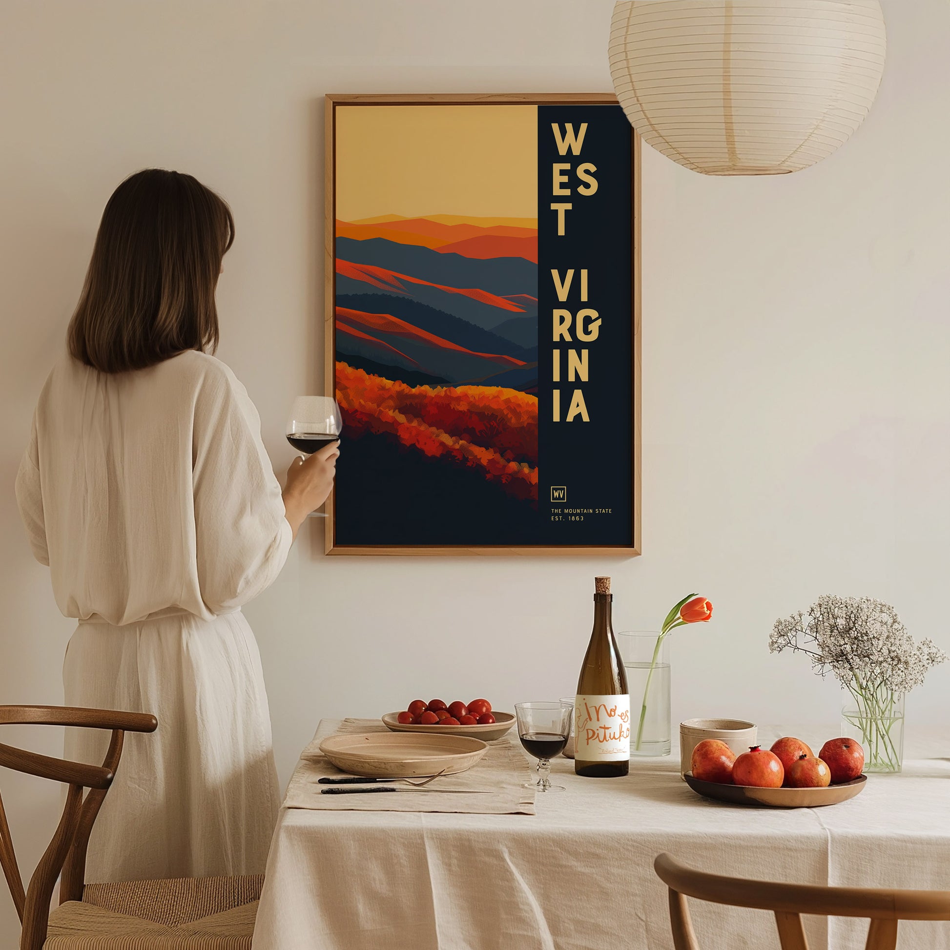 A woman in a white dress holds a glass of wine, admiring a West Virginia poster with vibrant autumnal colors. A table with fruit and wine is in the foreground.
