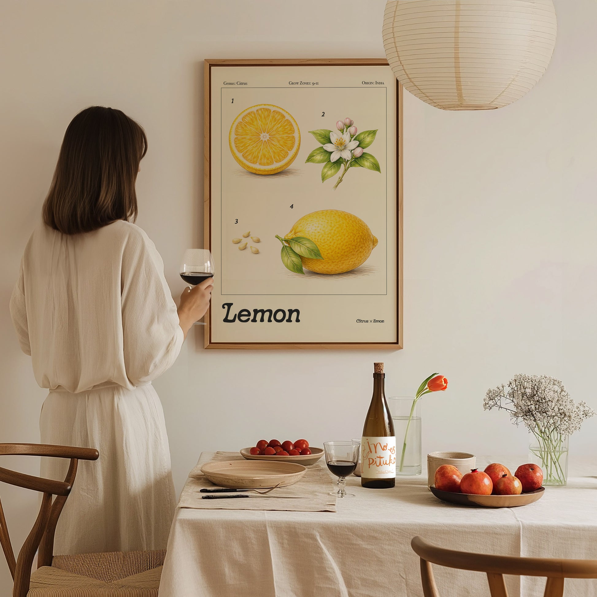 A woman in a white dress stands near a table, holding a glass of wine and admiring a framed botanical lemon poster on the wall.