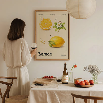 A woman in a white dress stands near a table, holding a glass of wine and admiring a framed botanical lemon poster on the wall.