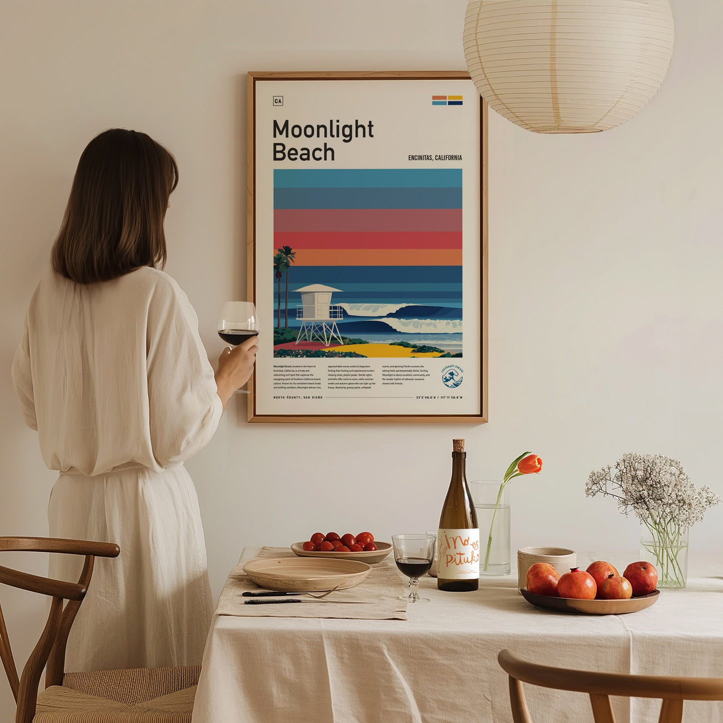 A woman in a white dress holds a glass of wine while standing next to a dining table set with fruit, a bottle, and a vase of flowers. Behind her, a framed poster titled Moonlight Beach features vibrant stripes and a coastal scene.
