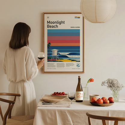 A woman in a white dress holds a glass of wine while standing next to a dining table set with fruit, a bottle, and a vase of flowers. Behind her, a framed poster titled Moonlight Beach features vibrant stripes and a coastal scene.