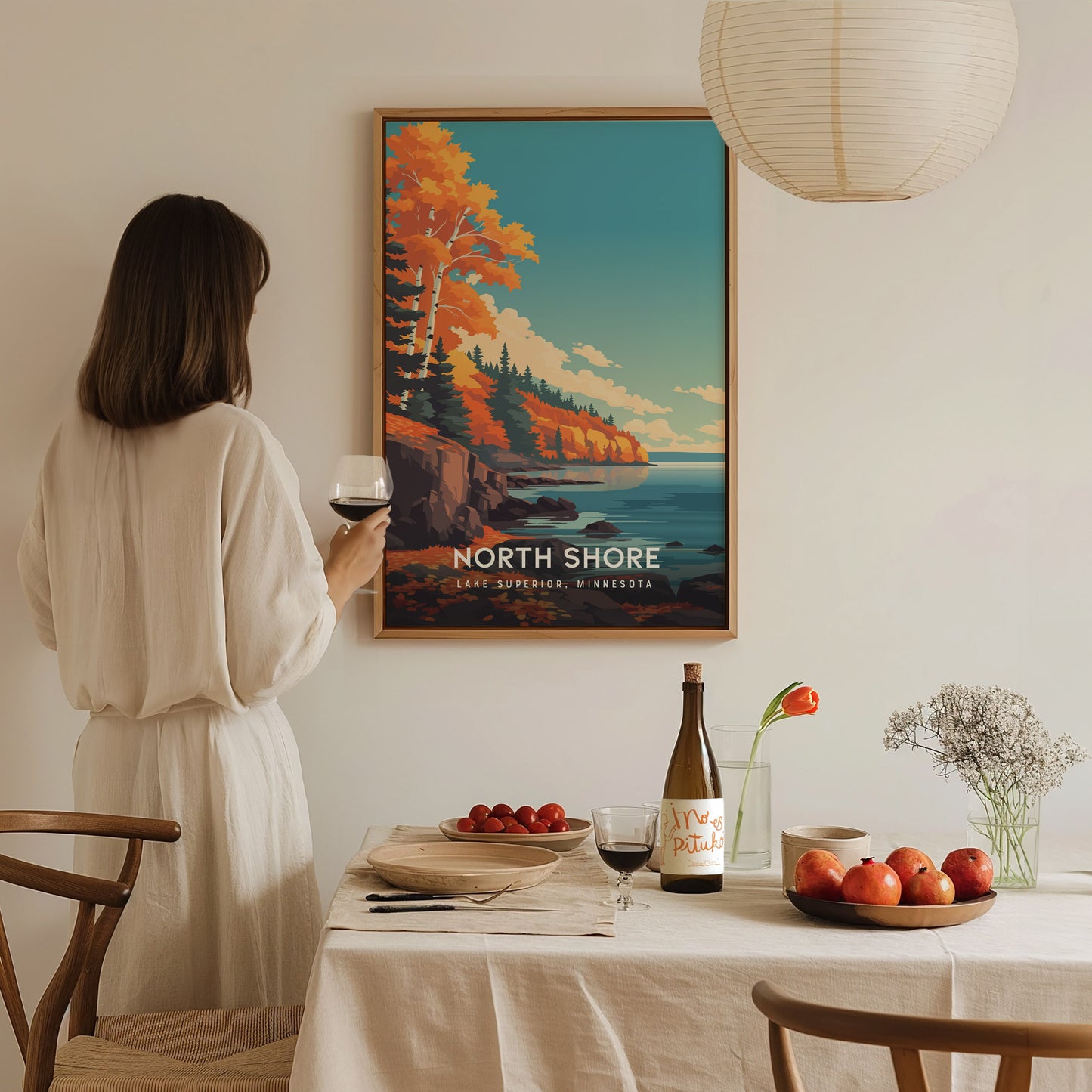 A woman in a white dress stands holding a glass of wine, gazing at a colorful poster of North Shore, Lake Superior, featuring vibrant autumn trees and a serene coastal scene.