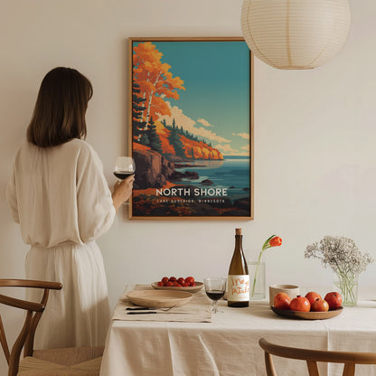A woman in a white dress stands holding a glass of wine, gazing at a colorful poster of North Shore, Lake Superior, featuring vibrant autumn trees and a serene coastal scene.