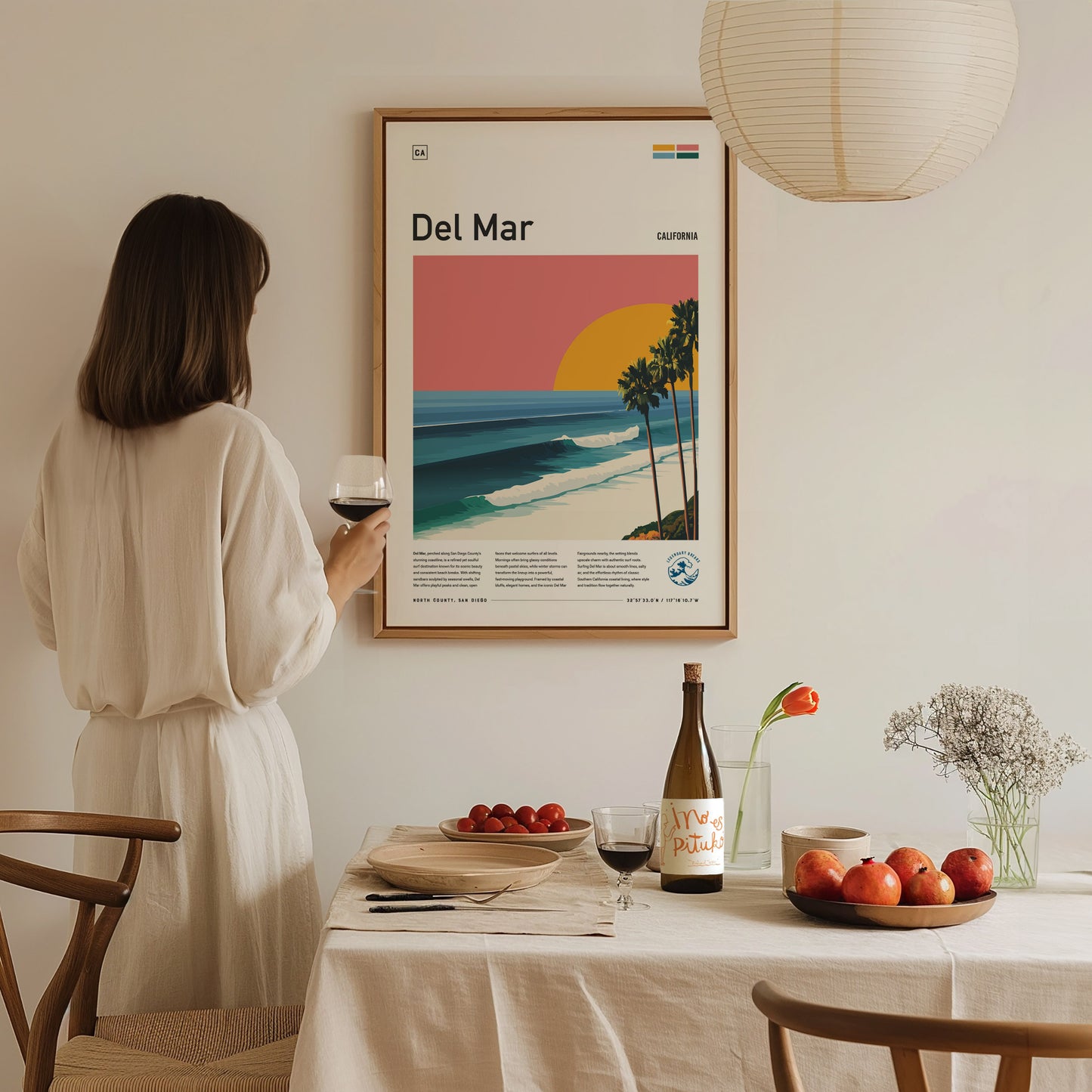 A woman in a light-colored dress stands holding a glass of wine, gazing at a framed poster of Del Mar Beach on a wall.