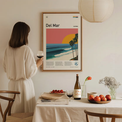 A woman in a light-colored dress stands holding a glass of wine, gazing at a framed poster of Del Mar Beach on a wall.