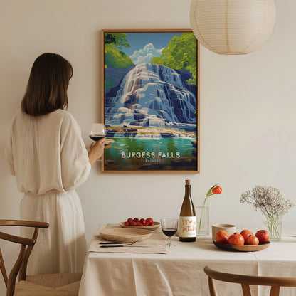 A person holding a wine glass admires a framed Burgess Falls poster. The room features a dining table set with fruit, wine, and a simple floral arrangement.