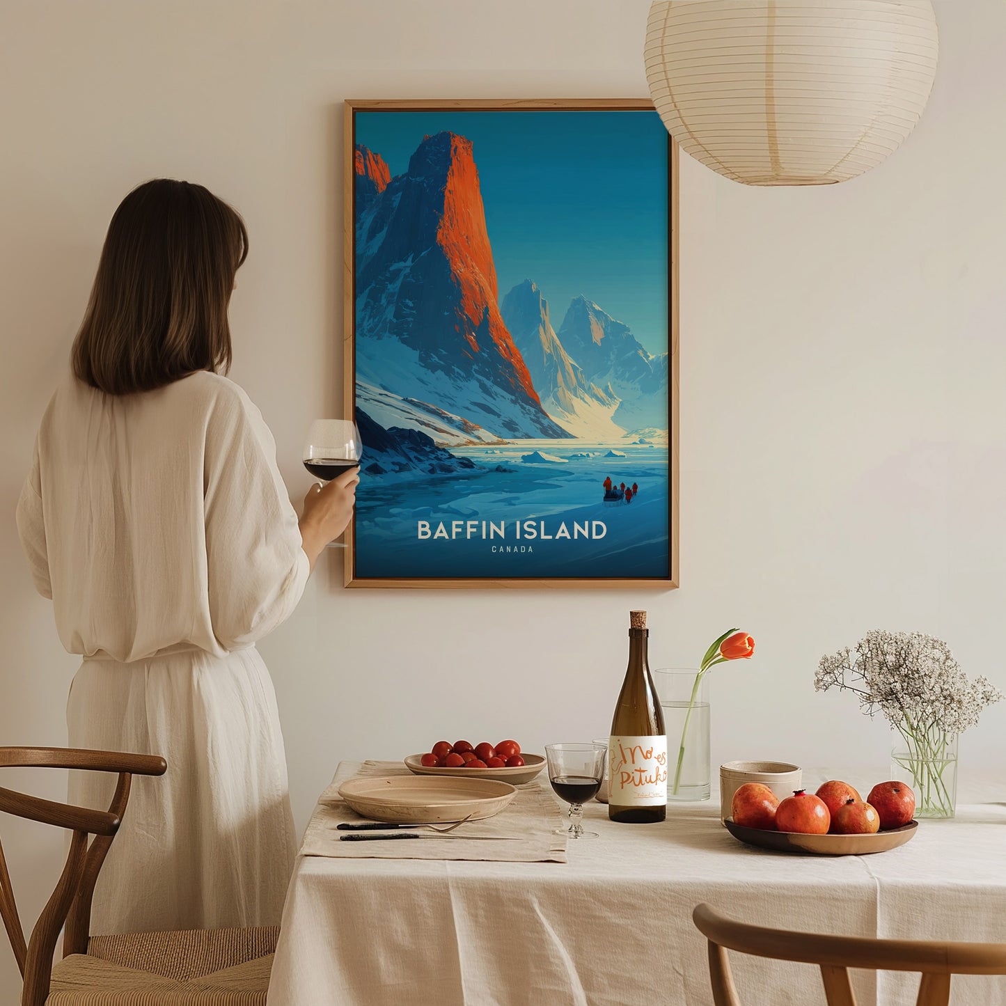 A woman in a cream-colored dress stands in front of a wall featuring a framed print of Baffin Island, showcasing vibrant mountains and a serene landscape.