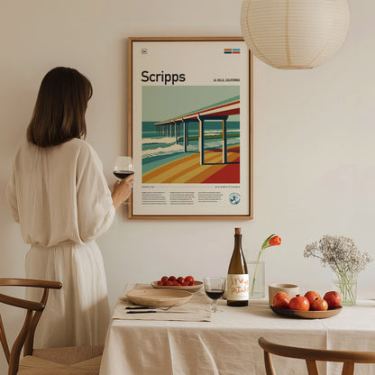 A woman in a white dress holds a glass of wine while admiring a framed Scripps Pier poster. The table is set with fruit, a wine bottle, and flowers.