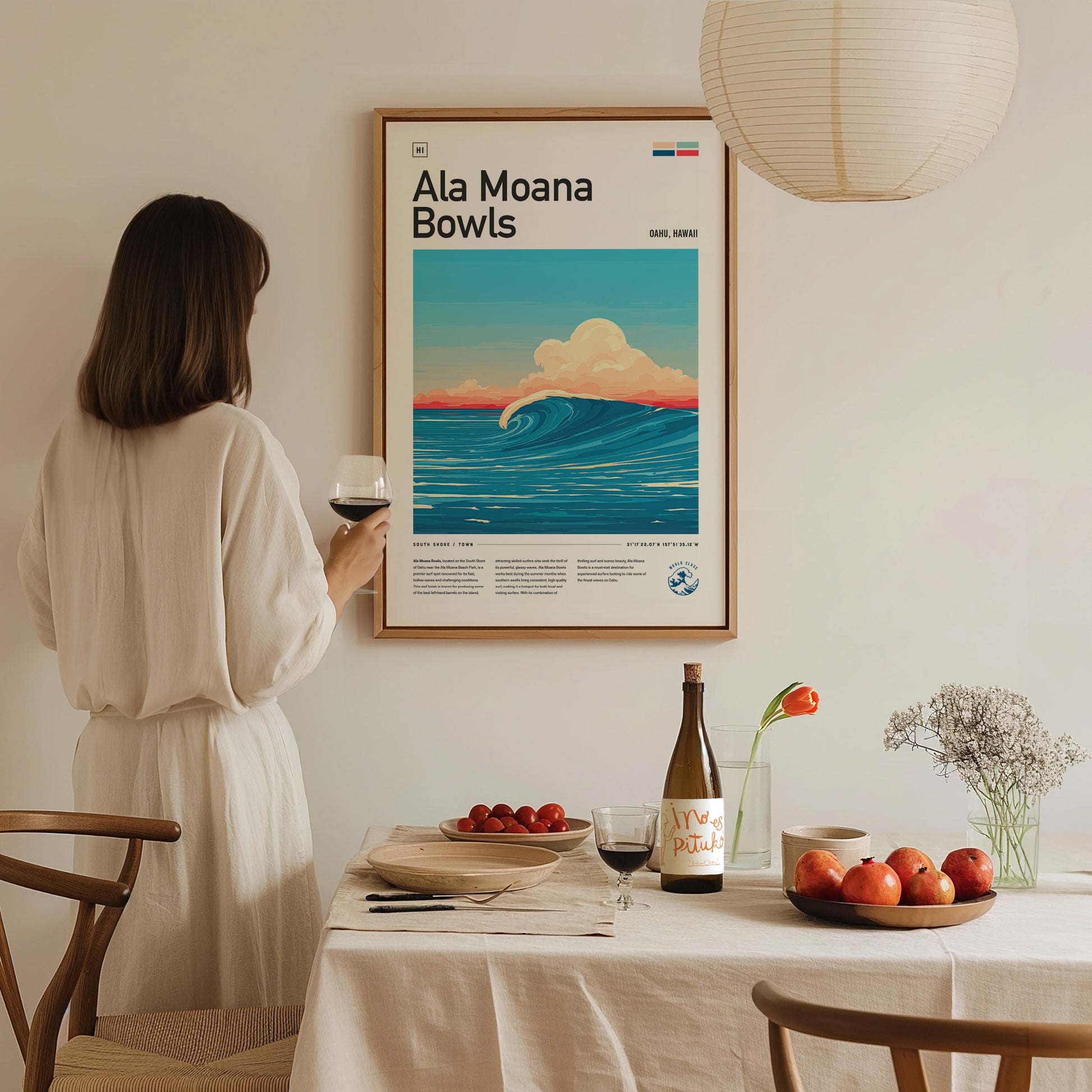 A woman in a white dress admires a framed Ala Moana Bowls surf poster on a wall. A table with fruit, wine, and flowers is in the foreground.